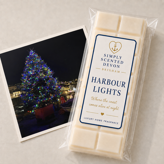 A packaged bar of Simply Scented Devon Brixham 'Harbour Lights' wax melts lies on a light neutral surface next to a printed photo of a lit Christmas tree at night. The wax bar is in clear plastic packaging with a white label featuring navy blue borders and gold text.