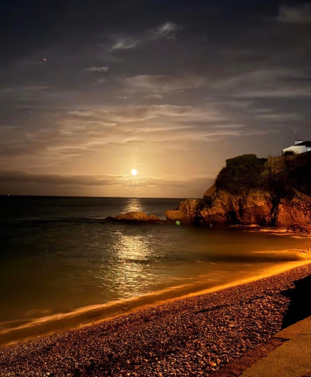 A scenic coastal landscape at night with a low glowing moon over calm water, rocky cliffs, a pebble beach in the foreground, and a dark cloudy sky.