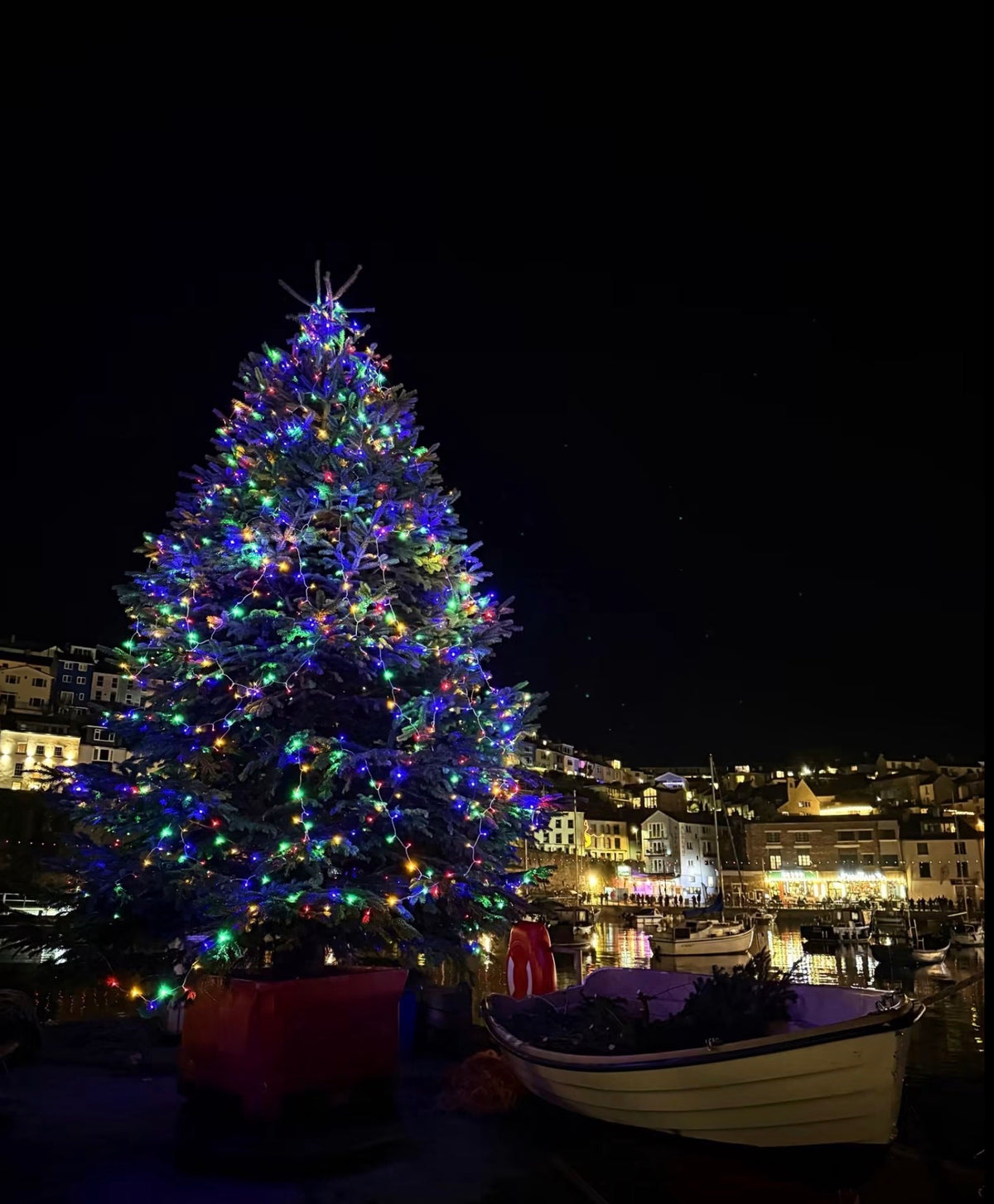 A large Christmas tree at night, decorated with colorful lights and ornaments, standing in Brixham harbor area with boats and illuminated buildings in the background.