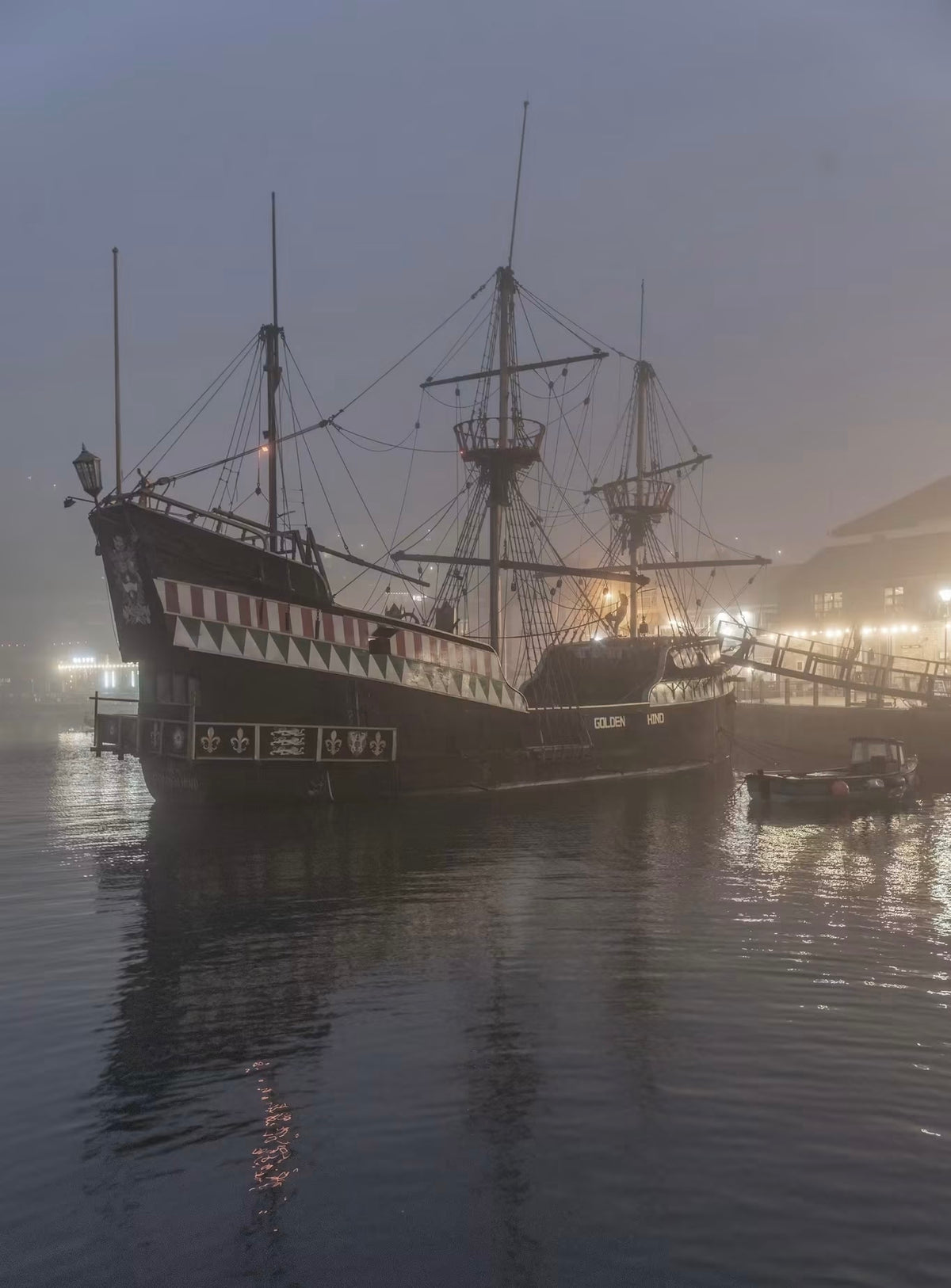 The Golden Hind, Brixham is a large historical sailing ship with three masts is docked in calm water at dusk, surrounded by mist and warm lights from the harbor. The ship has a dark hull with red-and-white striped rigging and white lettering on the side.